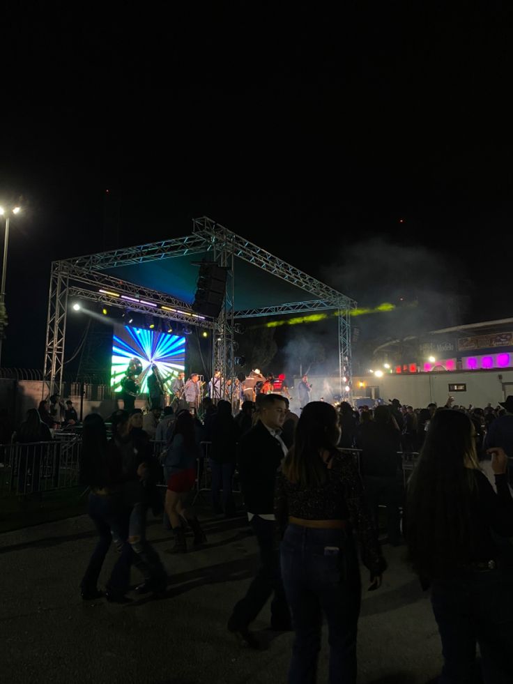 a group of people standing around in front of a stage with lights on it at night