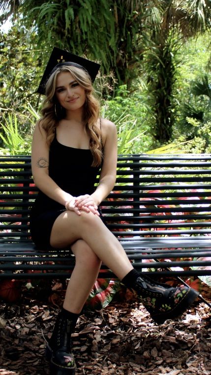 a woman sitting on top of a wooden bench in a park wearing black boots and a graduation cap