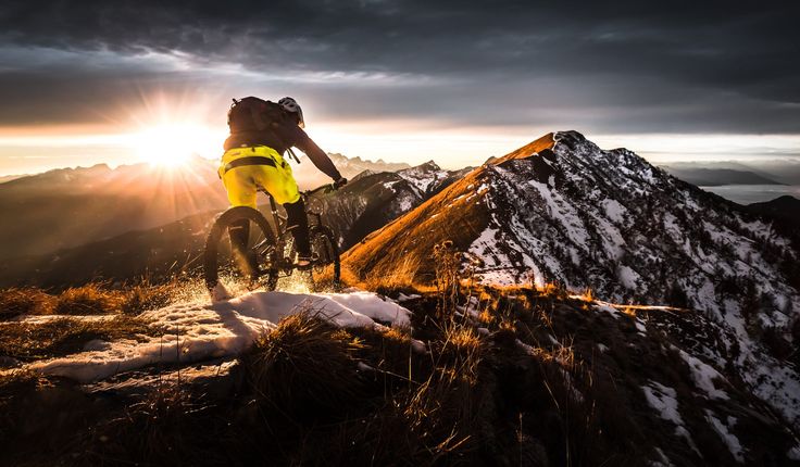 a man riding a mountain bike on top of a snow covered slope with the sun behind him