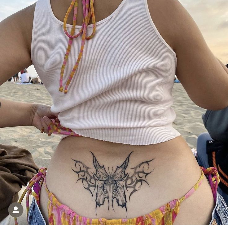 a woman with a butterfly tattoo on her stomach is sitting on the beach and looking at the camera