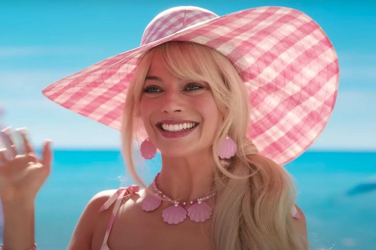 a woman with blonde hair wearing a pink hat and smiling at the camera while standing in front of the ocean