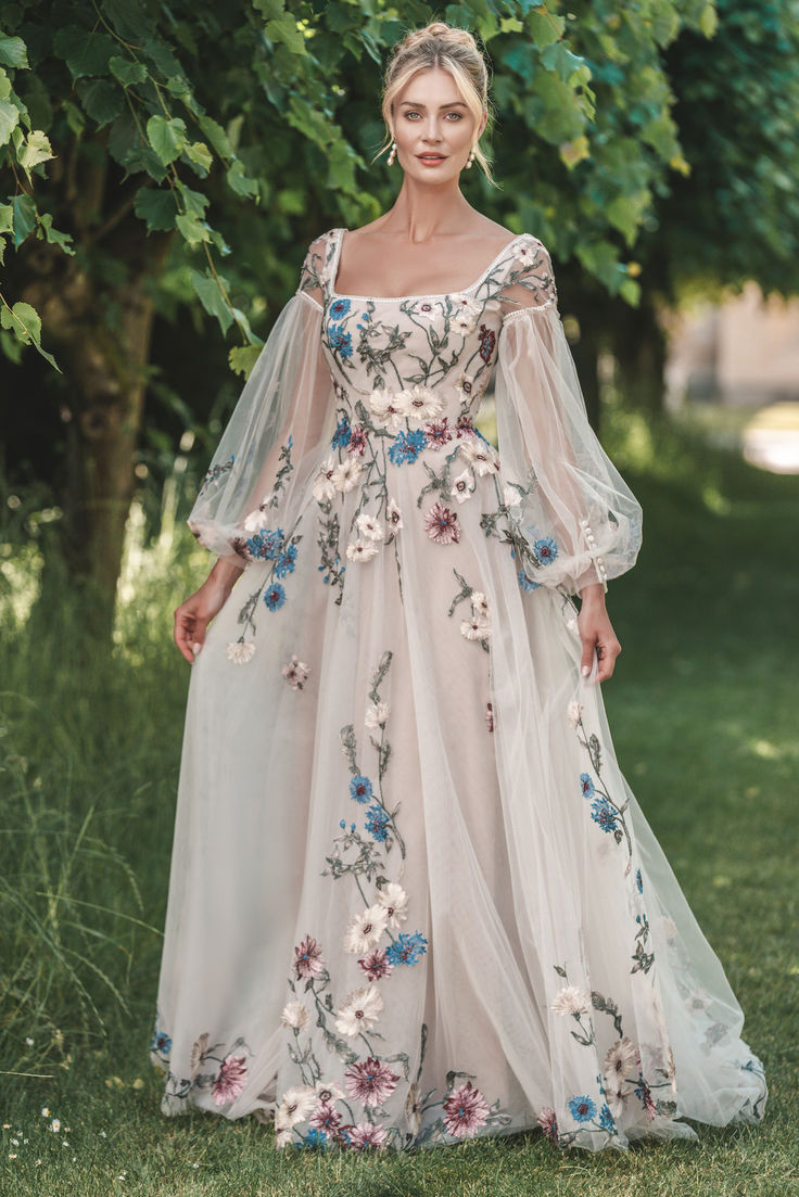 a woman in a white dress with flowers on it is standing near trees and grass