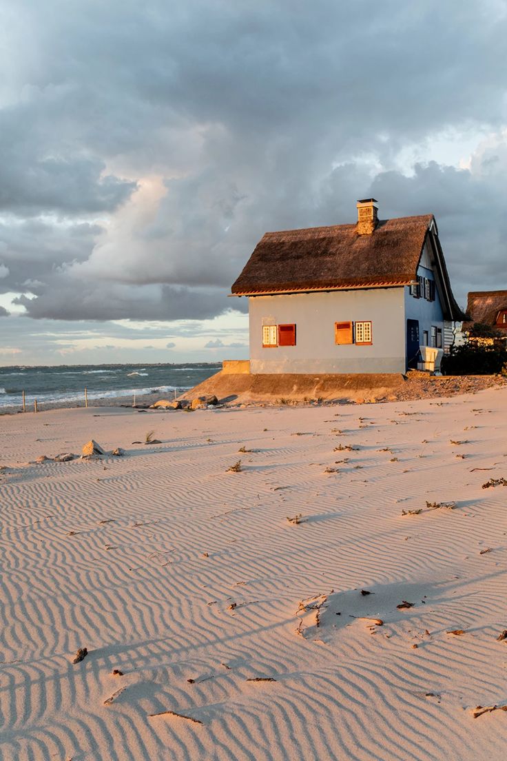 a house sitting on top of a sandy beach next to the ocean under a cloudy sky