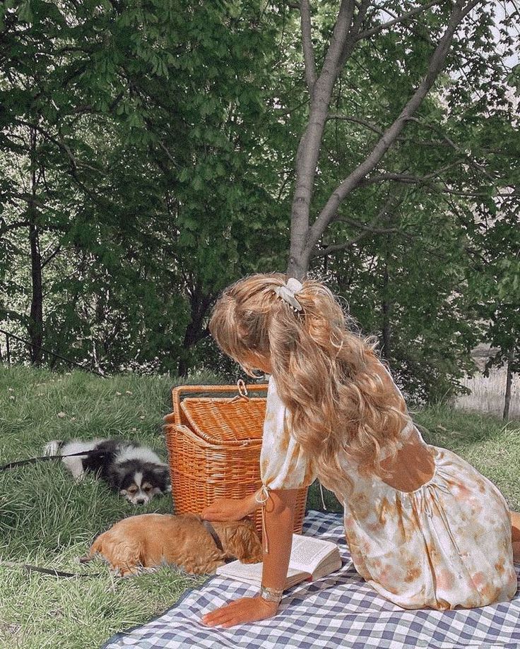 a woman sitting on top of a blanket next to a dog in the grass near a basket