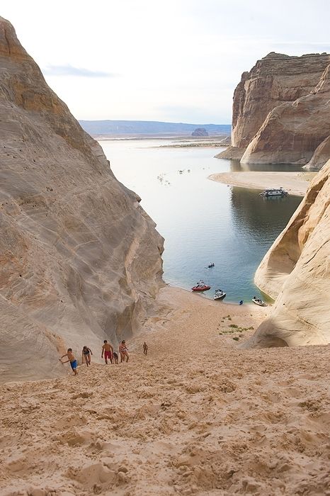 several people are walking on the sand near some water and cliffs, with boats in the distance