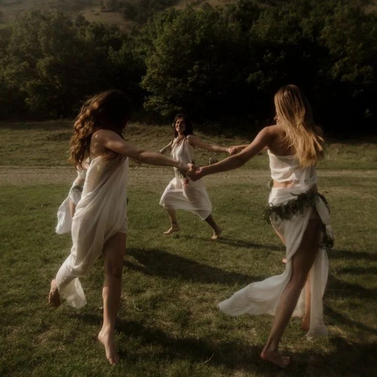 two women in white dresses holding hands and running across the grass with trees in the background