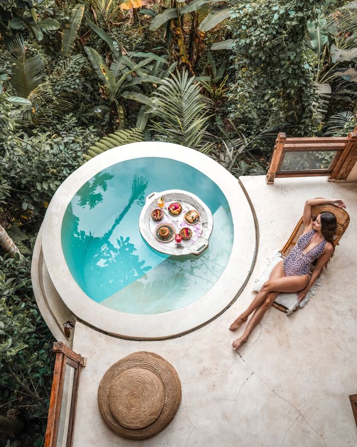 a woman sitting at a table next to a pool in the middle of some trees