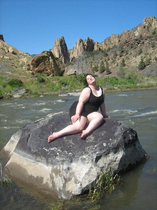 a woman sitting on top of a rock in the middle of a river with mountains in the background