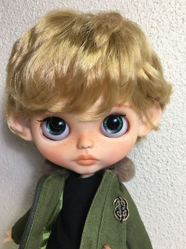 a close up of a doll with blonde hair and blue eyes wearing a green jacket