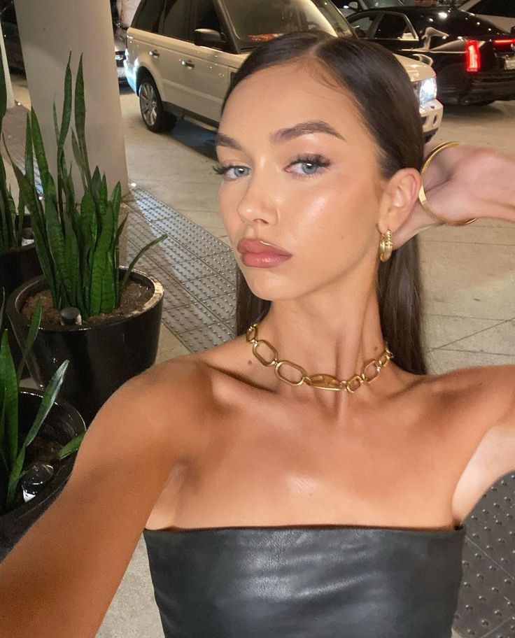 a woman wearing a black leather top and gold necklace sitting in front of a potted plant