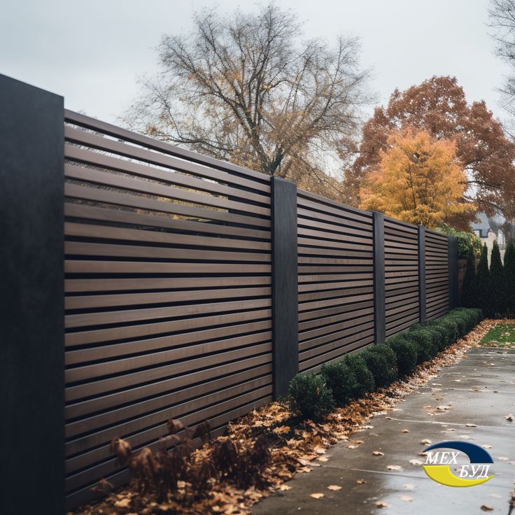 a wooden fence in the middle of a sidewalk with leaves on the ground and trees behind it