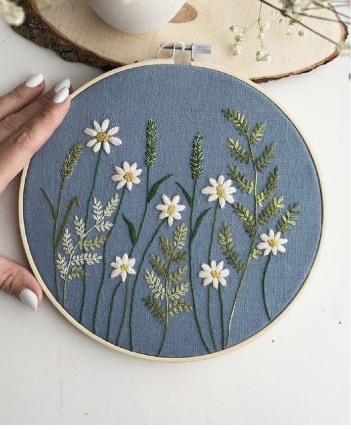 a woman is holding up a cross stitch embroidery project with daisies and grass on it