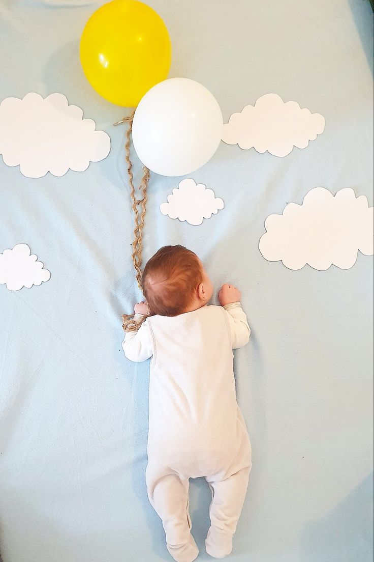 a baby laying on its back next to some balloons and clouds in the sky,