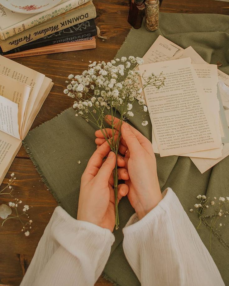 a person holding flowers on top of a wooden table next to books and other items