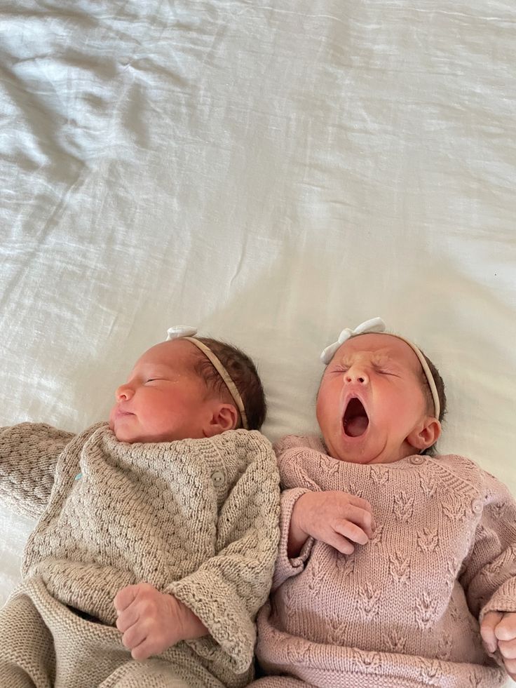 two baby babies laying next to each other on a white sheet covered bed, one yawning and the other yawning