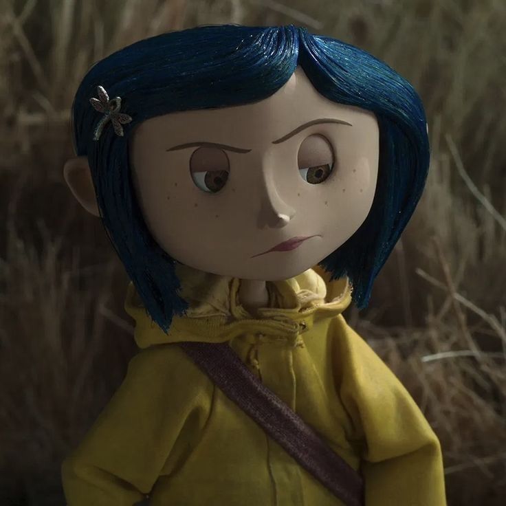 a doll with blue hair wearing a yellow jacket and standing in front of dry grass