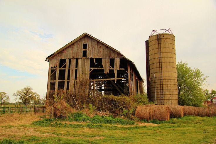 an old barn with hay bales in the foreground and a large silo behind it