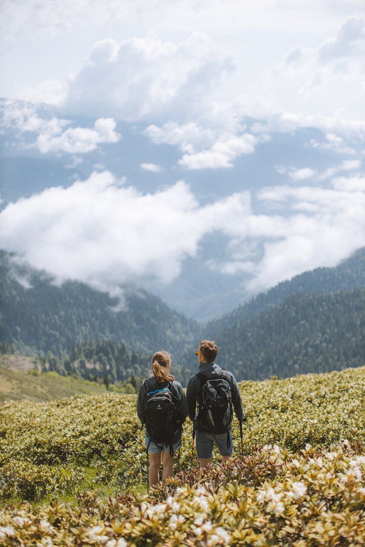 two people with backpacks standing in the middle of a field looking at mountains and clouds