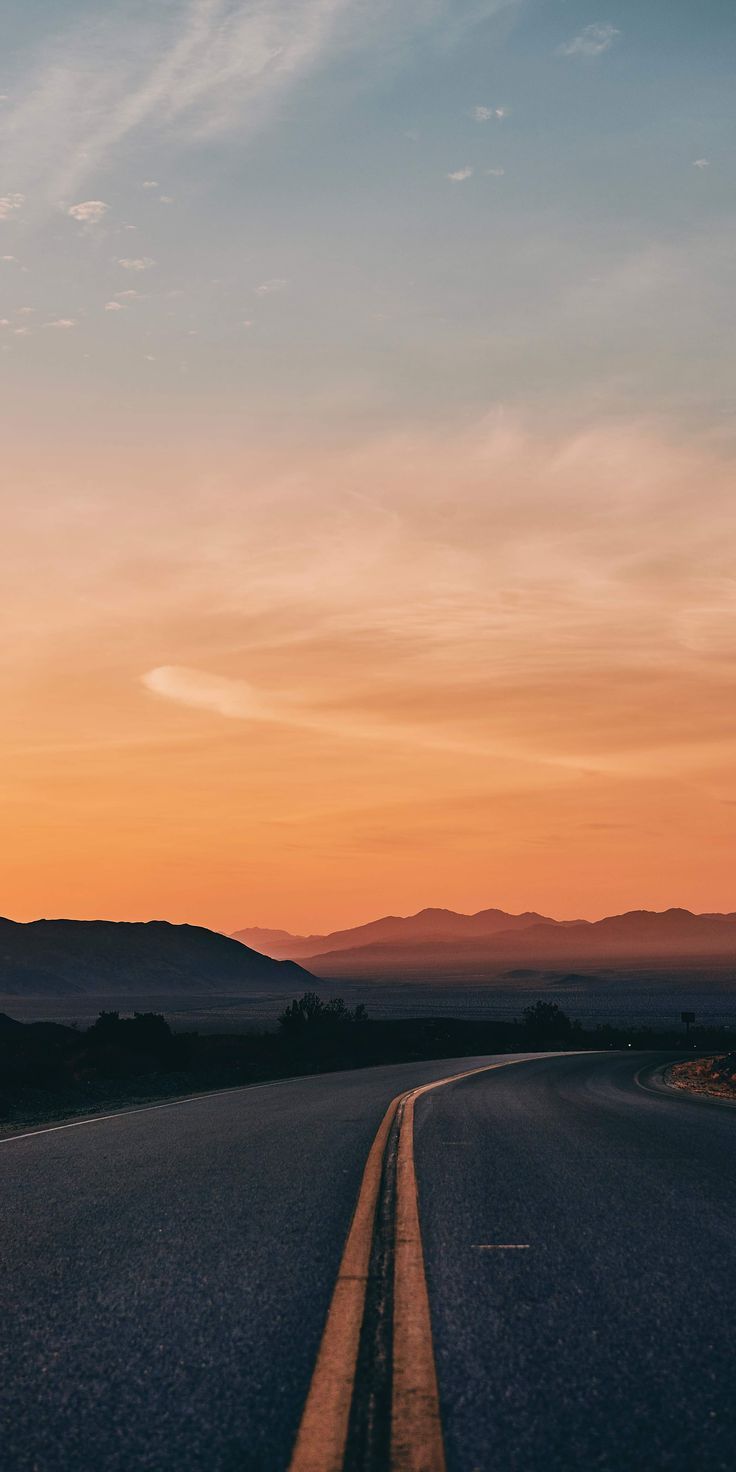 the sun is setting on an empty road with mountains in the distance and blue skies