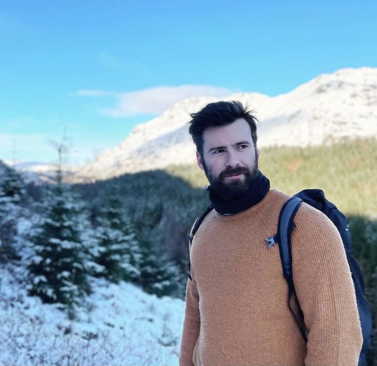 a man with a beard standing in the snow wearing a brown sweater and black pants