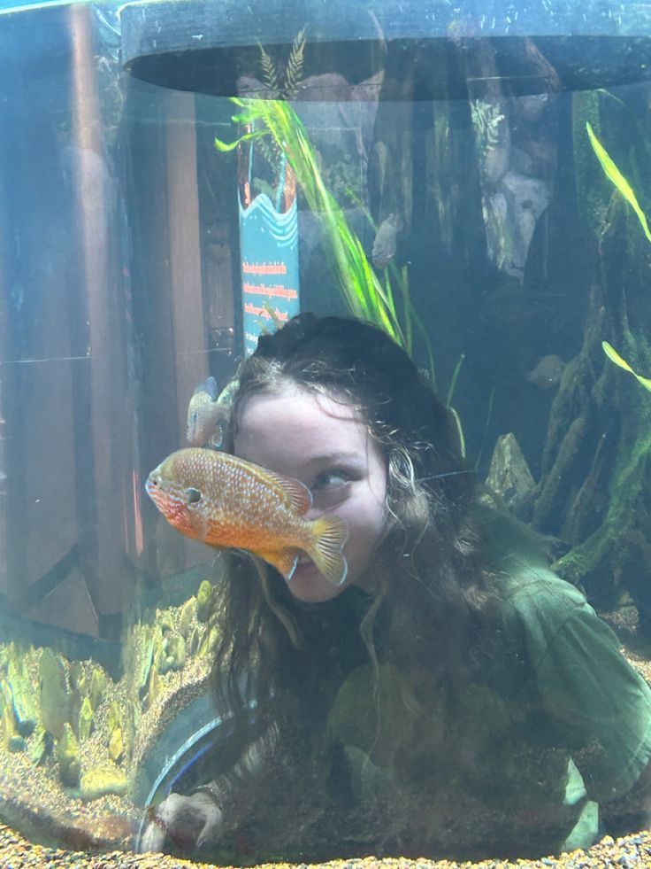 a young woman is looking at a fish in an aquarium with plants and rocks around her