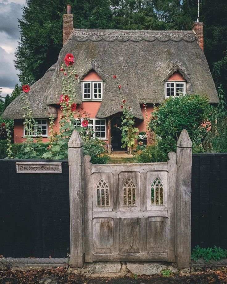 an old house with a thatched roof and flowers growing on the front gate, surrounded by greenery