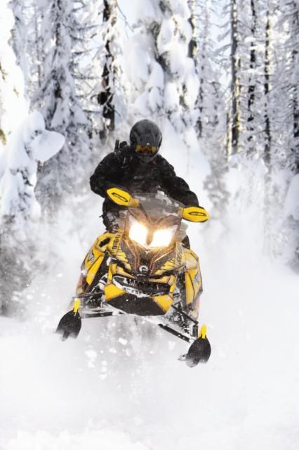 a man riding on the back of a yellow snowmobile down a snow covered forest