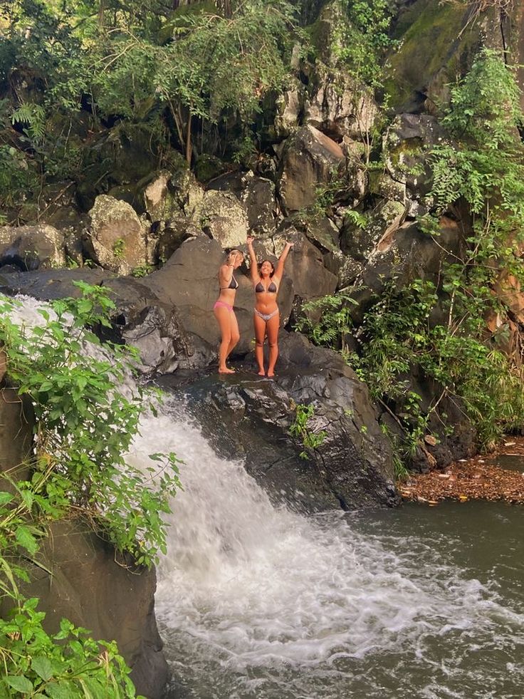 two women in bikinis standing on rocks next to a waterfall and holding their hands up