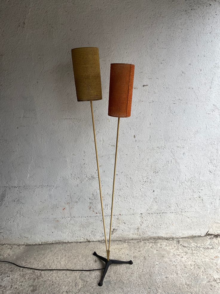 two floor lamps sitting next to each other on top of a cement floor in front of a white wall