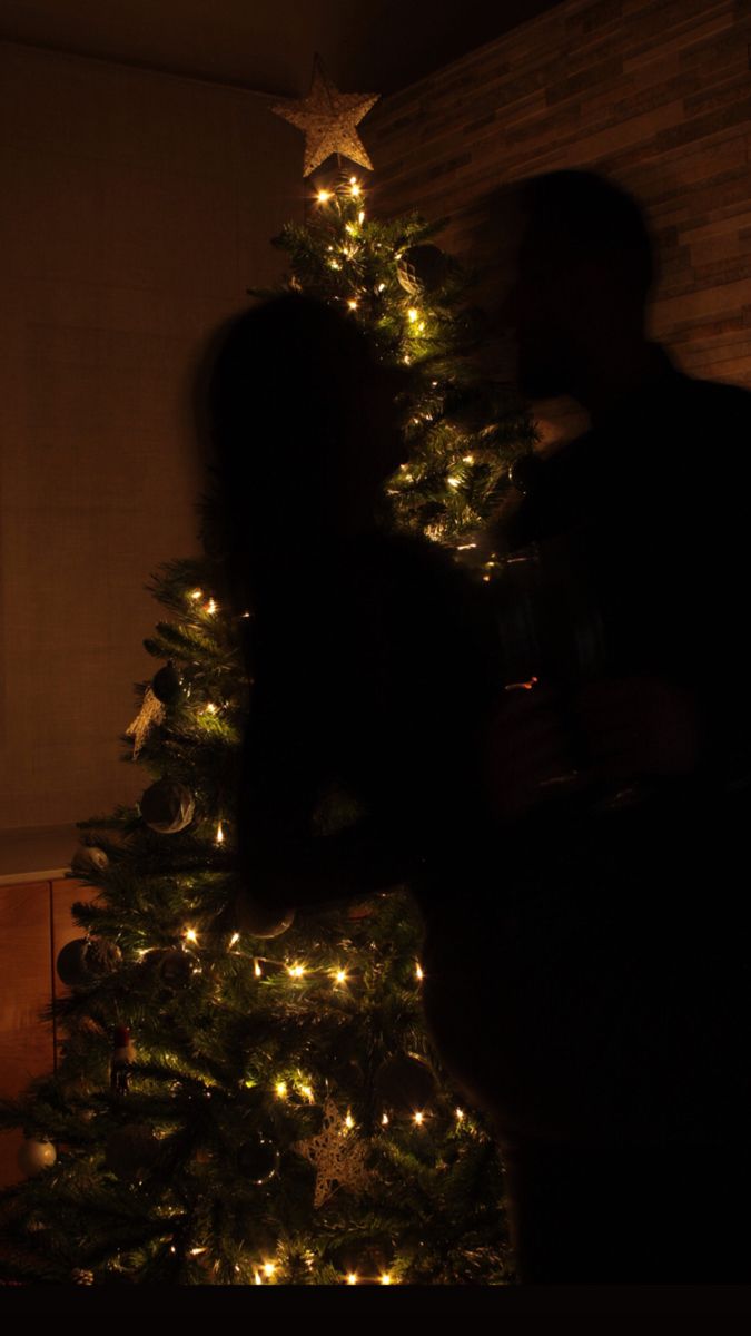 a man and woman standing next to a christmas tree with lights on it in the dark