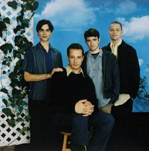 four men are posing for a photo in front of a blue sky and white fence