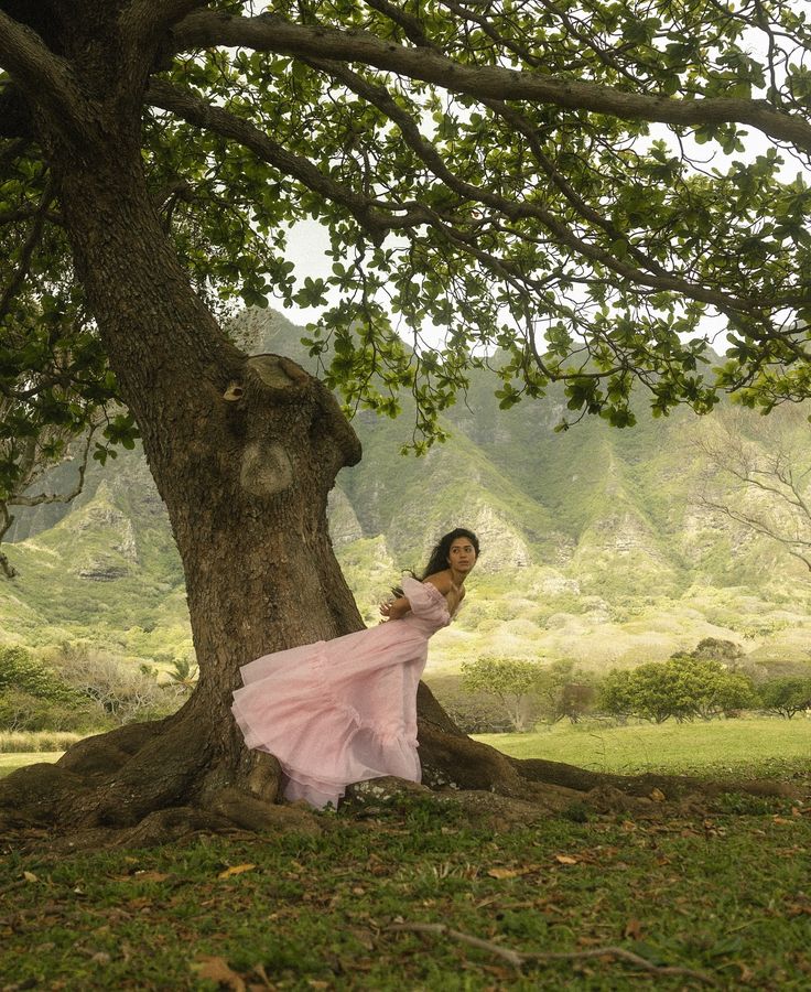 a woman in a long pink dress standing next to a tree with her arms around the trunk