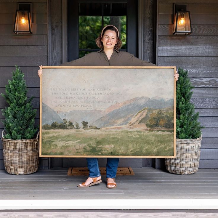 a woman holding up a large painting in front of a house with potted plants