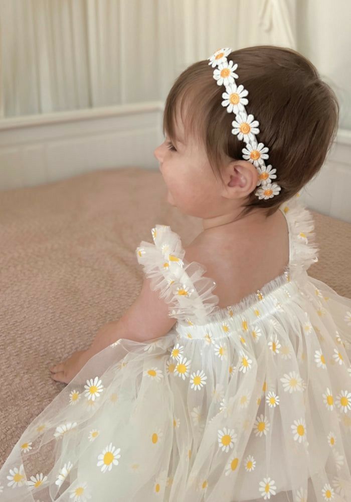 a baby sitting on the floor wearing a white dress with daisies in it's hair