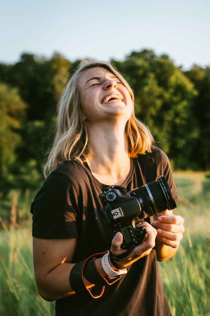 a woman holding a camera up to her face in the grass with trees in the background