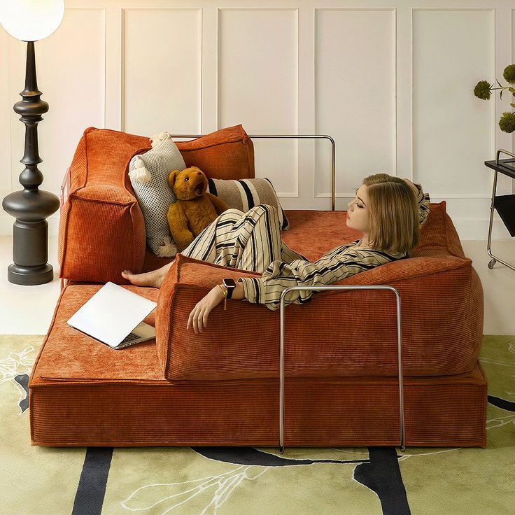 a woman laying on top of an orange couch next to a lamp and teddy bear