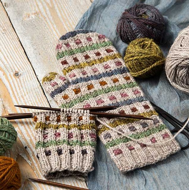 knitted mitts and yarn on a wooden table with knitting needles, balls of yarn and crochet hooks