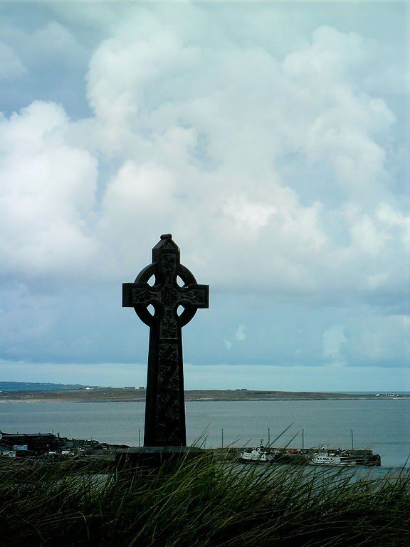 a large cross sitting on top of a lush green field next to the ocean with clouds in the sky