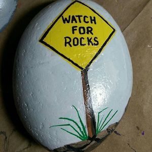 a painted rock with a yellow sign that says watch for rocks on it and grass growing underneath