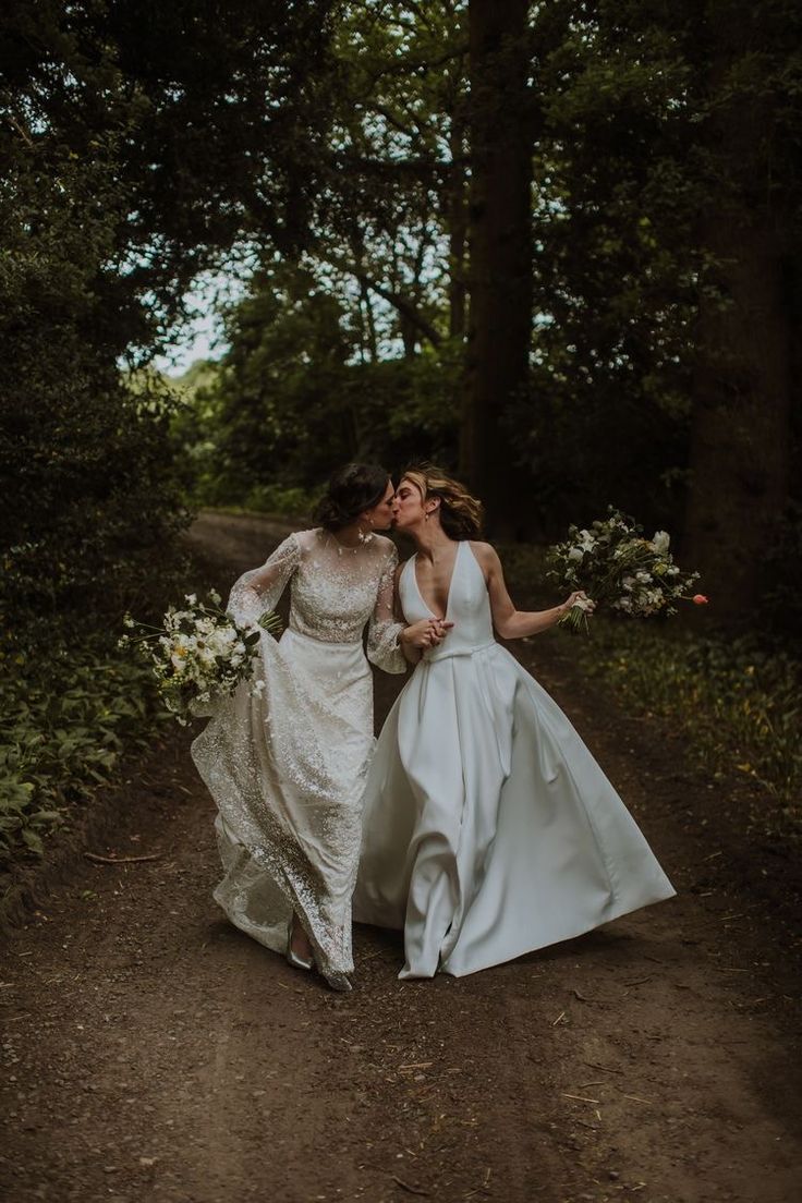 two brides walking down a dirt road in the woods with their arms around each other