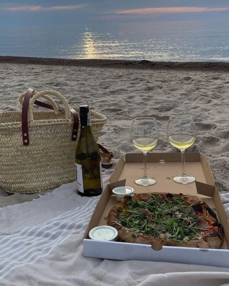 there is a pizza and two glasses of wine on the sand at the beach,