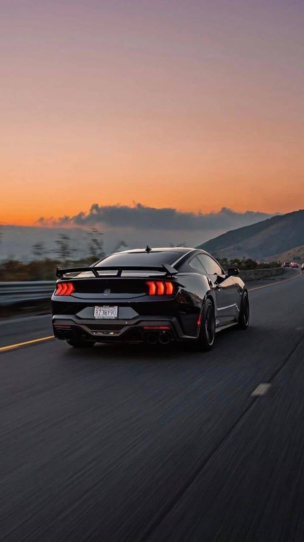 the rear end of a black sports car driving down a highway at sunset with mountains in the background