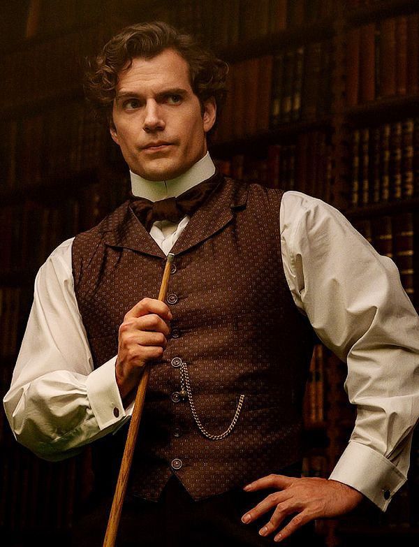 a man in a vest and bow tie holding a cane with bookshelves behind him