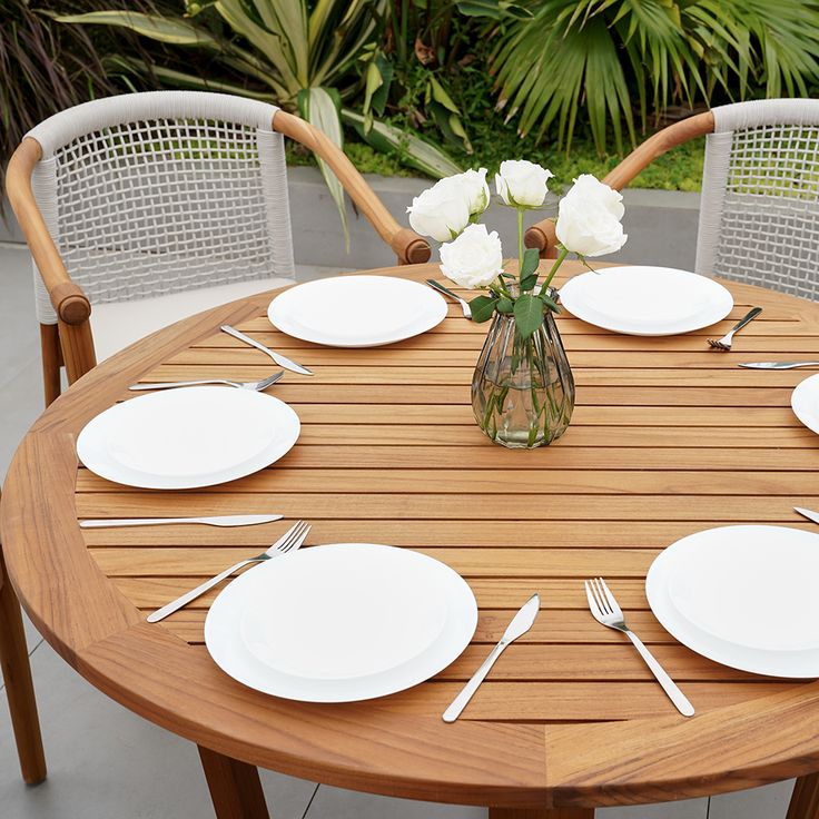 a wooden table with white plates and silverware on it, in front of palm trees