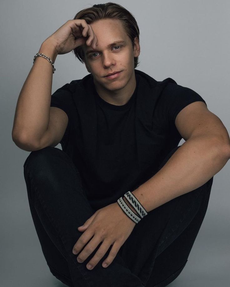 a young man sitting on the ground with his hands behind his head and looking at the camera