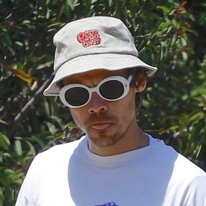 a man wearing sunglasses and a hat is standing in front of some trees with his hand on his hip