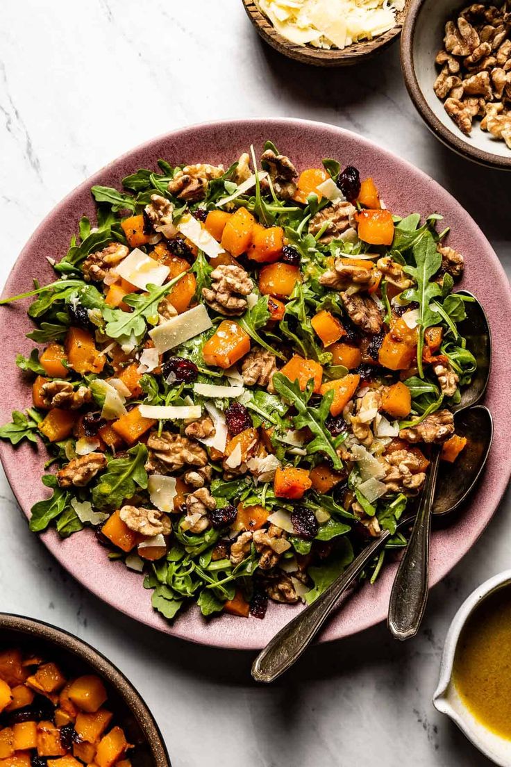 a pink plate topped with a salad next to bowls of vegetables and dressing on a white table