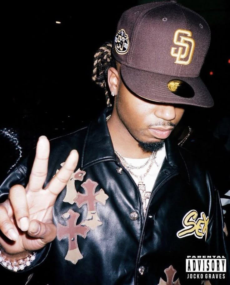 a man in a leather jacket and baseball cap making the peace sign with his hand