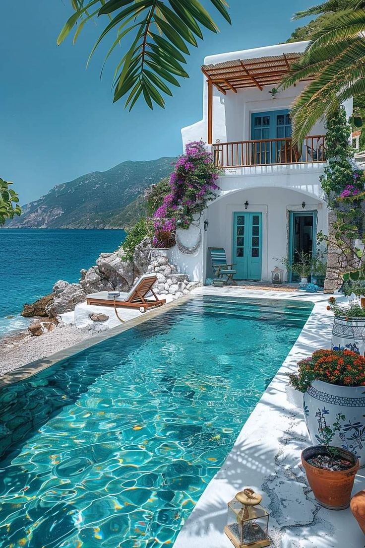 an outdoor swimming pool next to a white house with blue shutters and flowers in pots