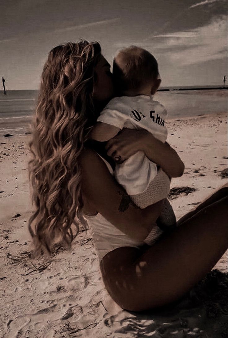 a woman sitting on top of a sandy beach next to the ocean holding a baby